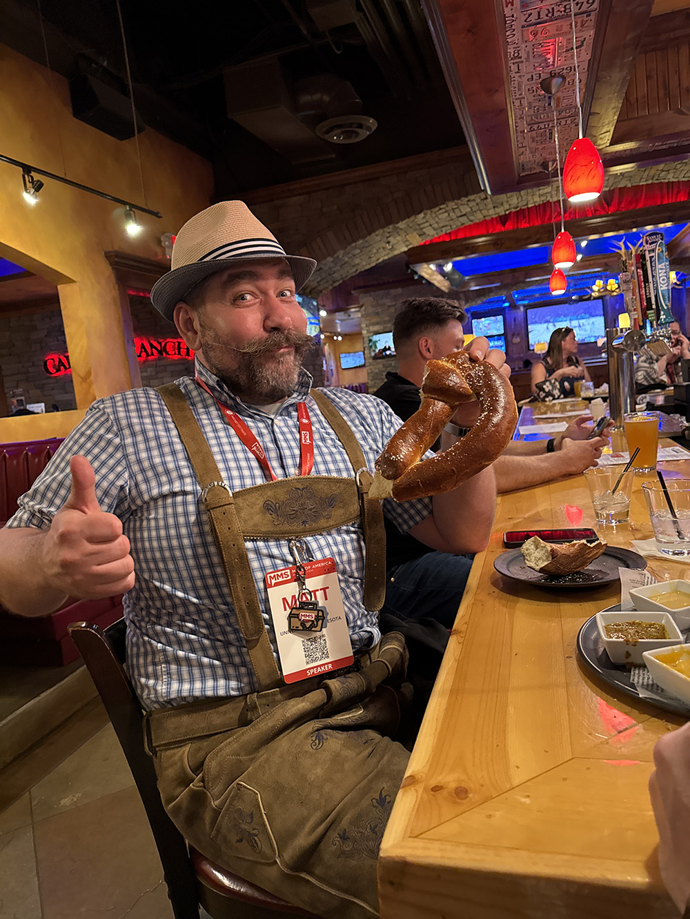 Matt partakes of a giant pretzel while wearing lederhosen seated at a bar.